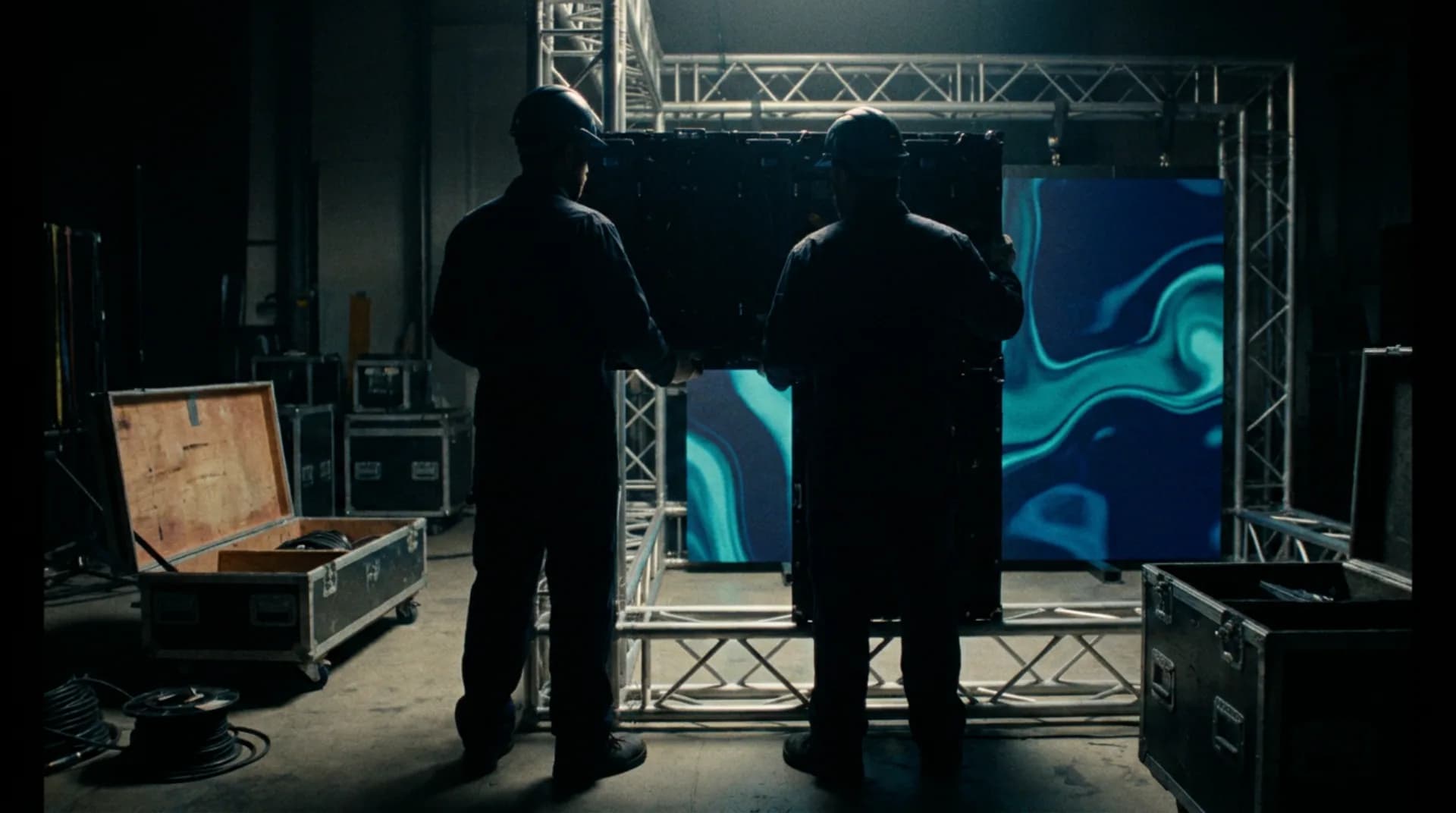 Two technicians latching LED cabinets onto a truss, lit from behind by the panels they have already hung